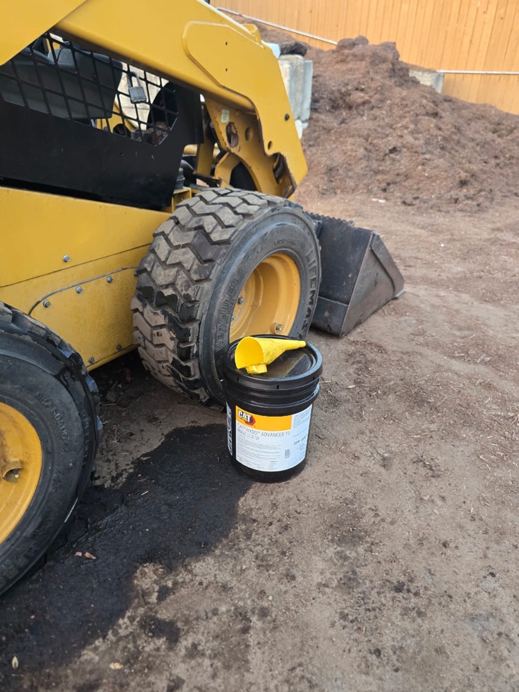 Bucket of Cat hydraulic oil with a funnel next to the front tire, ready to refill
