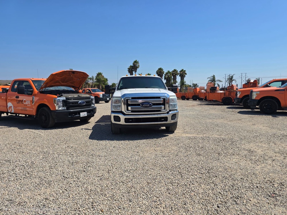 White Ford truck parked between orange service trucks, with utility trailers in the background